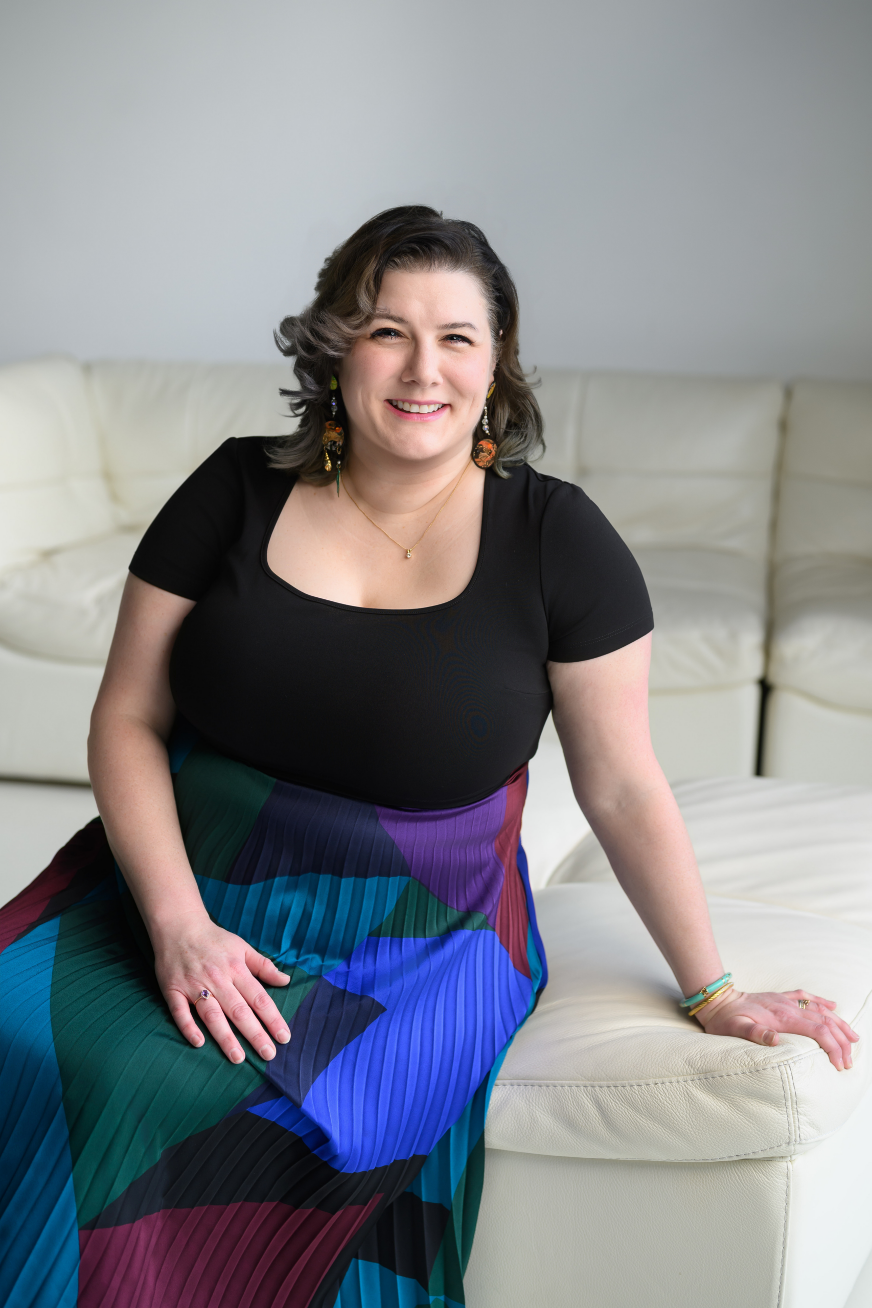 Oct 2026_Grace Headshot C. Grace Whiting, co-founder and principal of Whiting Communications, in a black dress and blue, purple, green skirt, sitting on a white couch